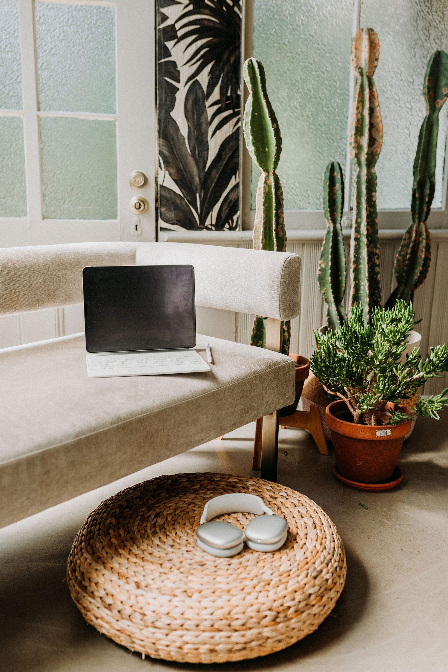 iPad attached to Magic Keyboard sitting on a modern couch next to cactus plants. In front of the couch is a wicker poof where Airpods Max are displayed.