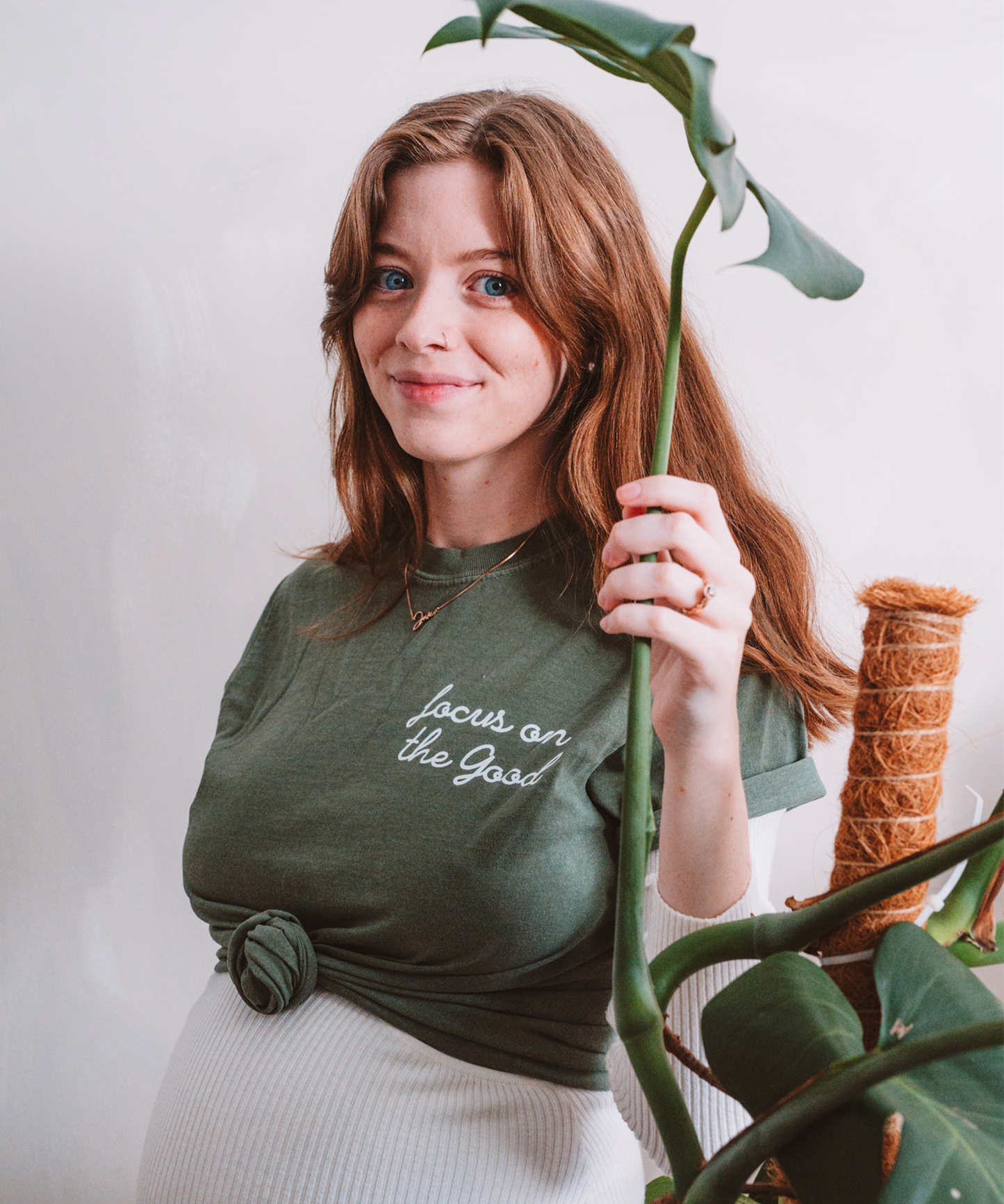 A model is smiling while holding a monstera leaf. She is wearing a "Focus on the Good" tea in moss that is stylishly knotted in the front.