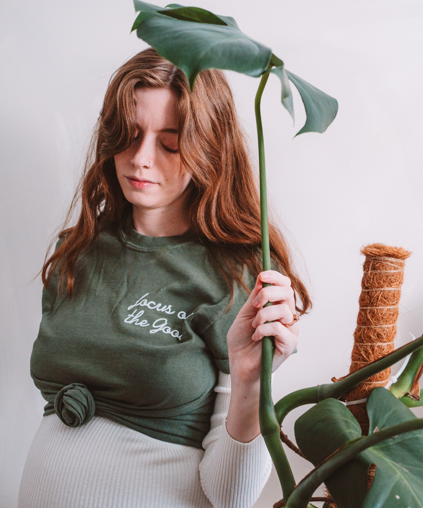 A model is posed holding a monstera leaf looking down at her shirt. She is wearing a "Focus on the Good: t-shirt in moss.