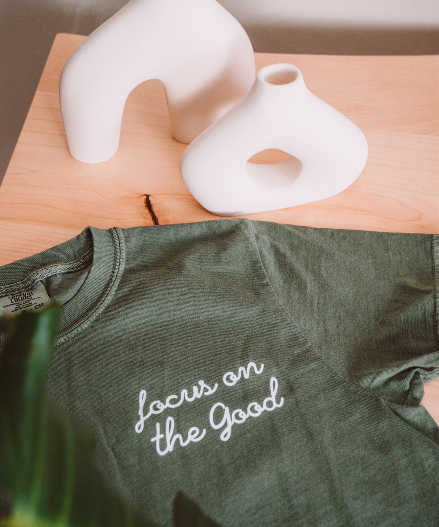 A "Focus on the Good" t-shirt is laying on a desk with two asymmetrical vases in the background.