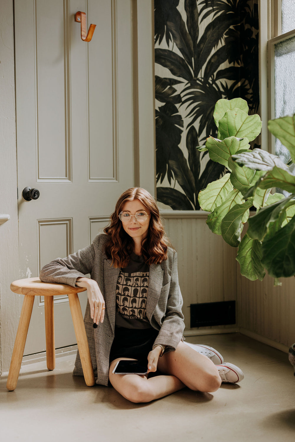 Kirstin from KDigitalStudio is sitting on the floor with the iPad mini in her lap. She leans one arm against a stool. Surrounding her are tons of plants. She's wearing vintage glasses, a blazer, and a Beatles t-shirt.