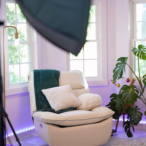 White armchair with green cushions in a room with large windows and a plant.