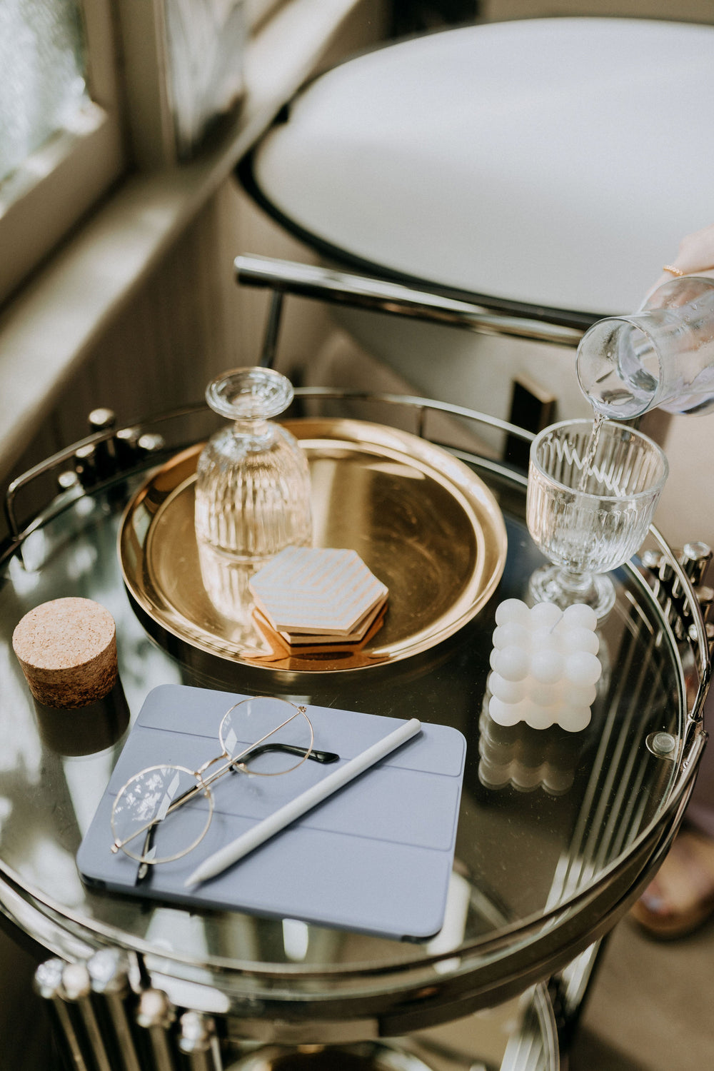 A purple iPad mini, an Apple Pencil, and vintage circular glasses are sitting on a modern bar cart. Off to the side, someone is pouring water into a glass.