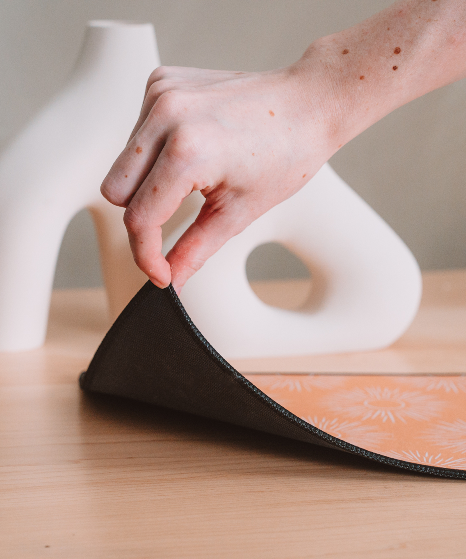 A hand is lifting up the corner of the Soleil mousepad to reveal the black, anti-slip rubber backing. In the background are two asymmetrical vases.