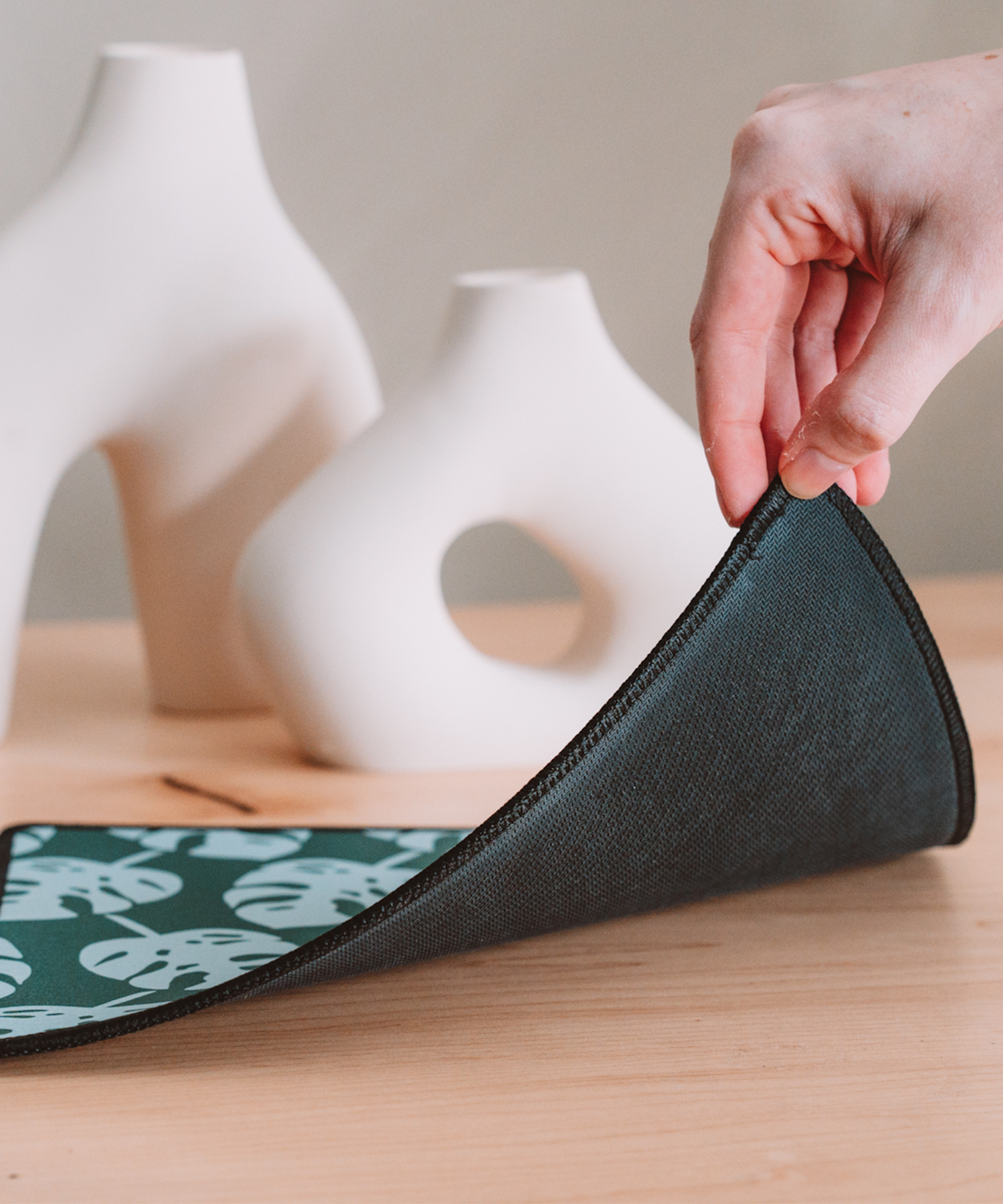 A hand is lifting a corner of the Monstera mousepad to reveal the black anti-slip rubber backing. In the background are two asymmetrical vases.