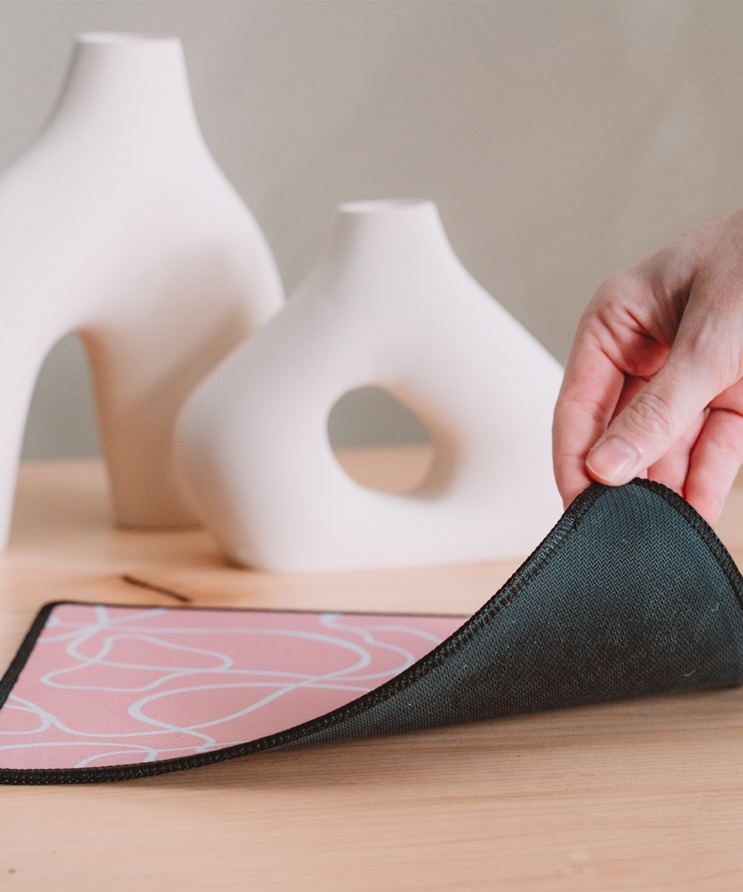 A hand is lifting up the corner of the Terrain mousepad to reveal the black, anti-slip rubber backing. In the background are two asymmetrical vases.