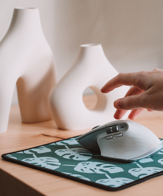 A hand is reaching for a mouse on top of the Monstera mousepad. In the background are two asymmetrical vases.