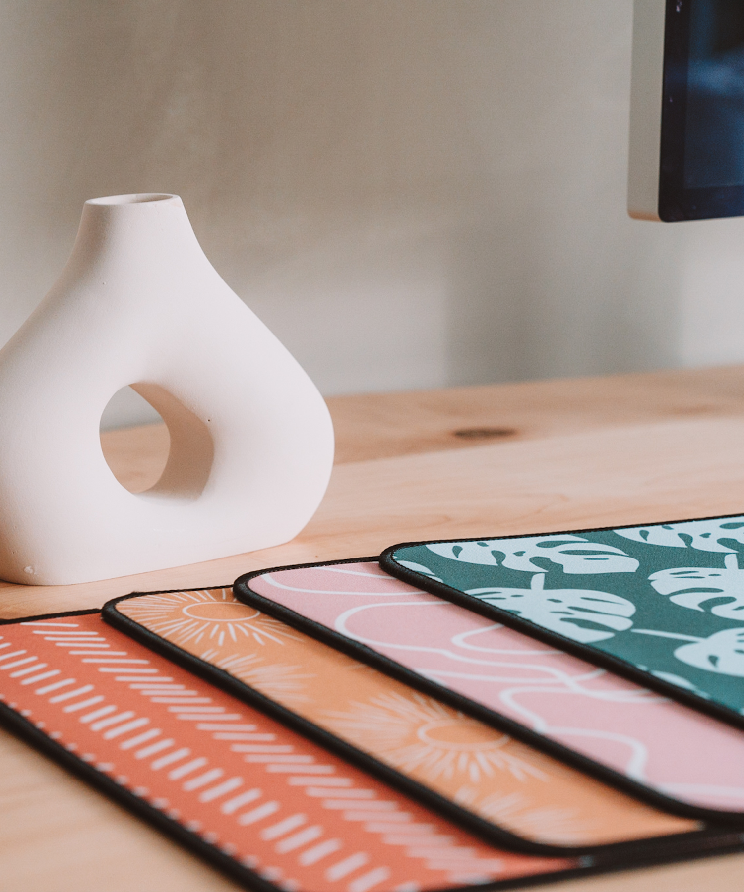 All mousepads are stacked laying on a desk with an asymmetrical vase in the background