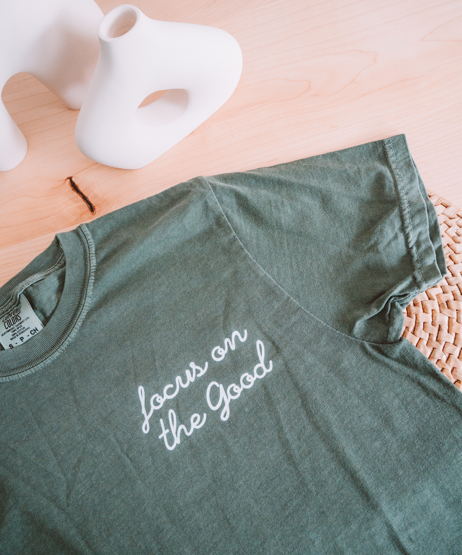 The "Focus on the Good" t-shirt in moss is laying on a desk with two asymmetrical vases in the background.