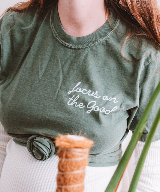 A closeup of the "Focus on the Good" t-shirt in Moss. The text is centered on the left breast. The shirt is stylishly knotted in the front.