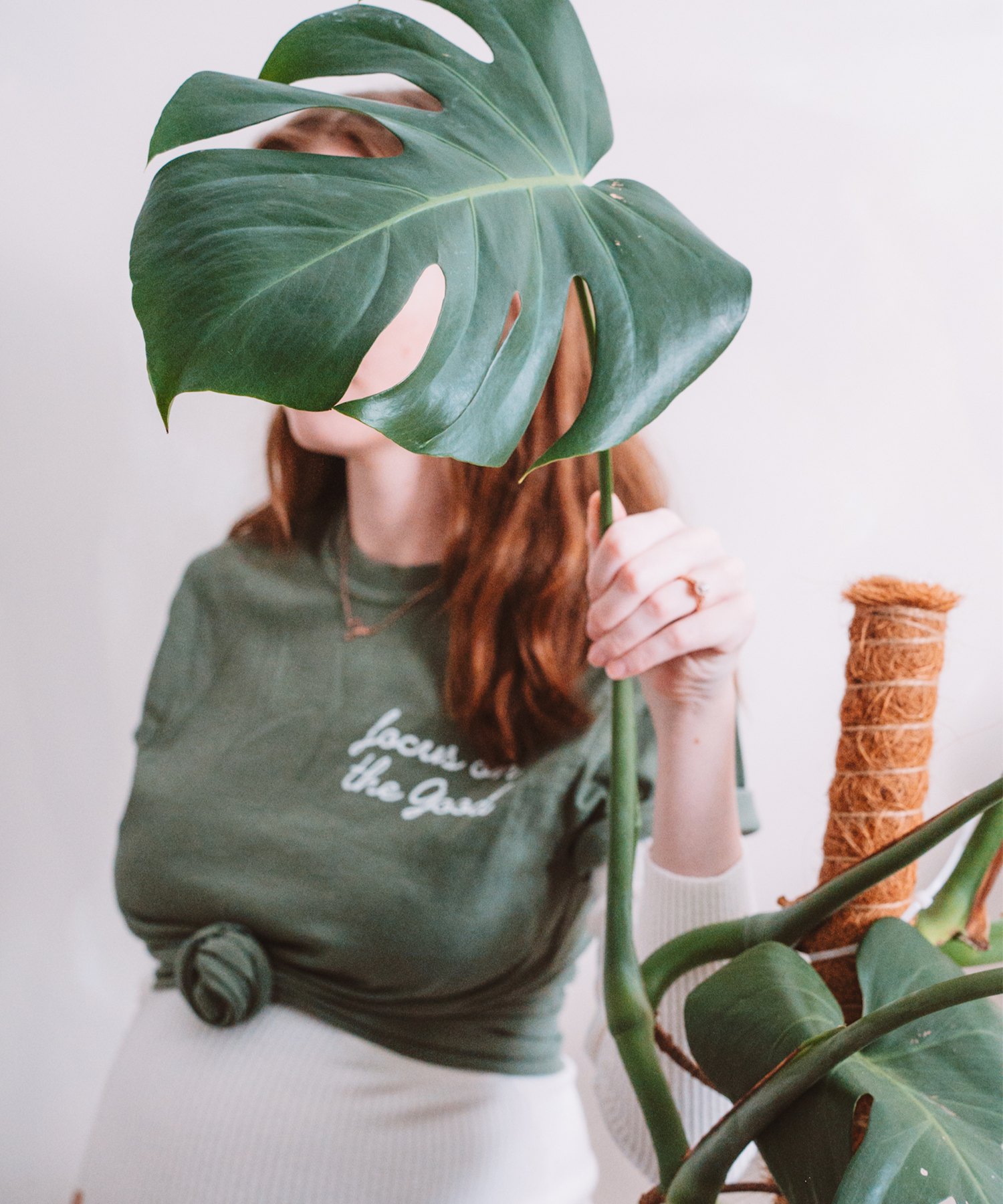 A model is posed hiding her face behind a large monstera leaf. She is wearing a "Focus on the Good" t-shirt, stylishly knotted in the front.
