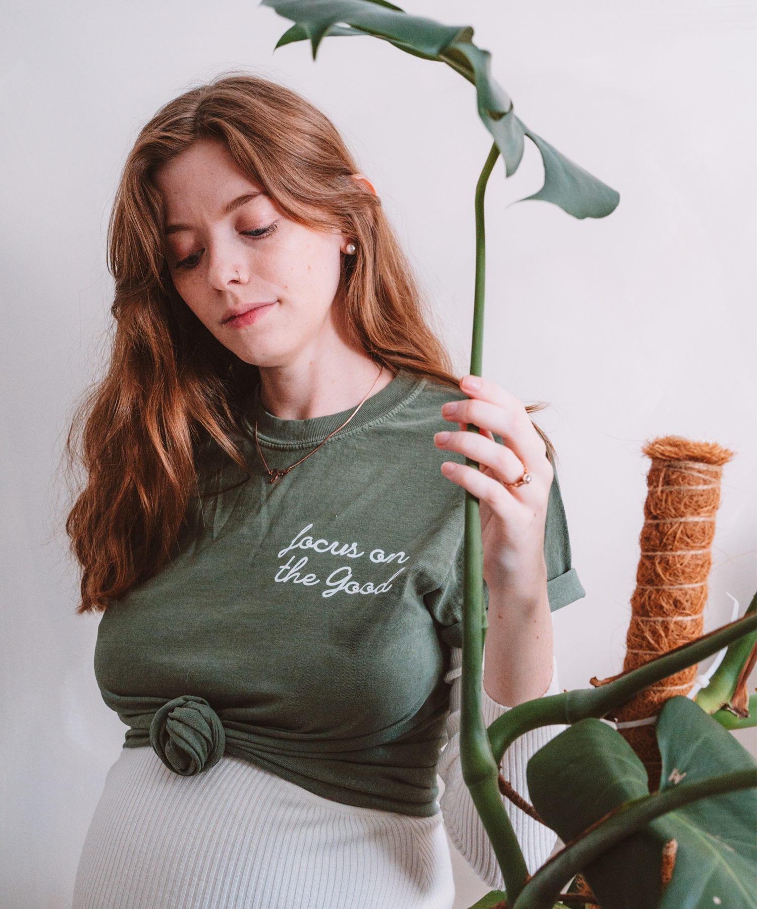 A model is posed holding a monstera leaf and looking down at her "Focus on the Good" t-shirt in moss
