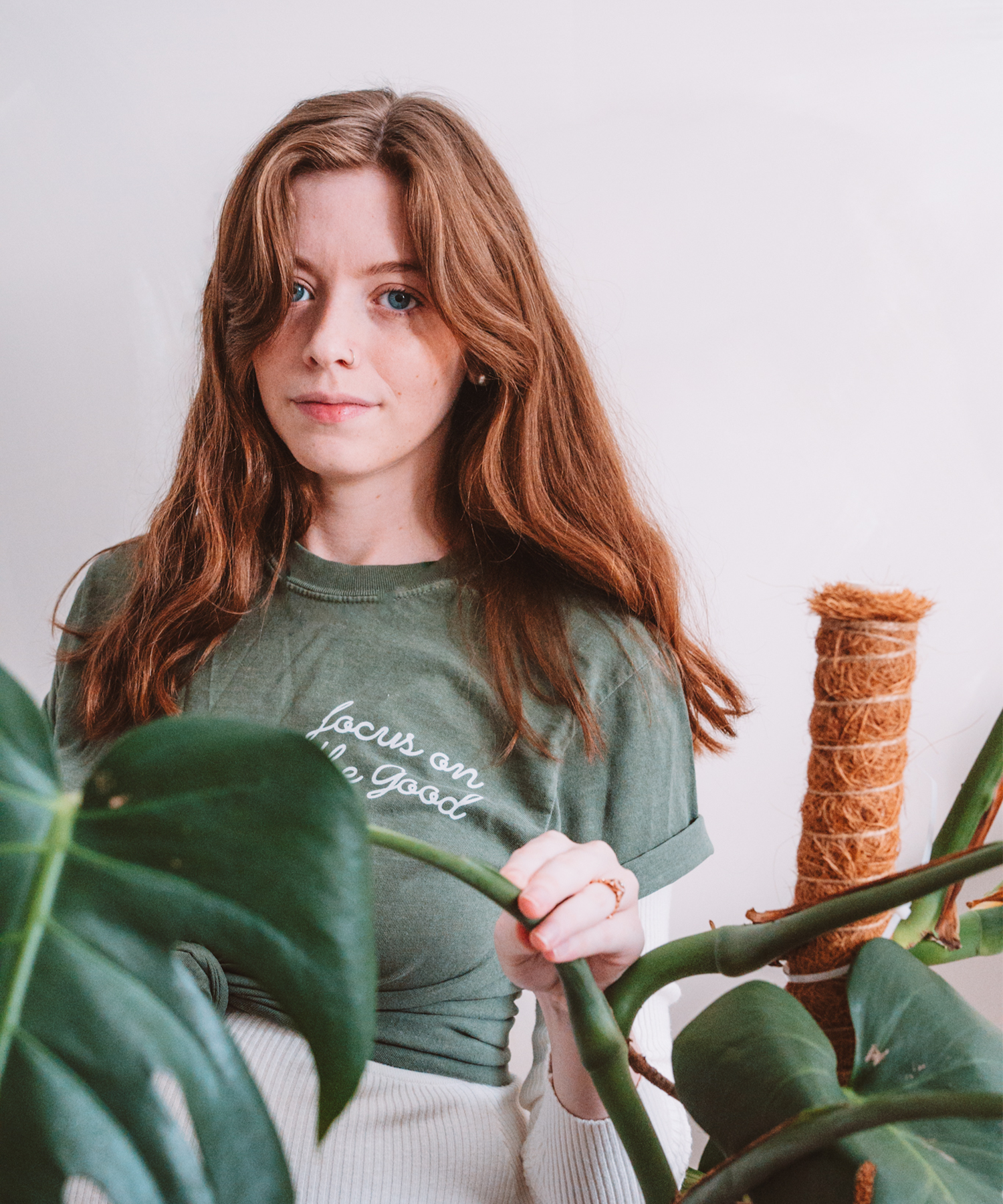 A model is posed behind a monstera plant. She is wearing the "Focus on the Good" t-shirt in moss, stylishly knotted in the front.