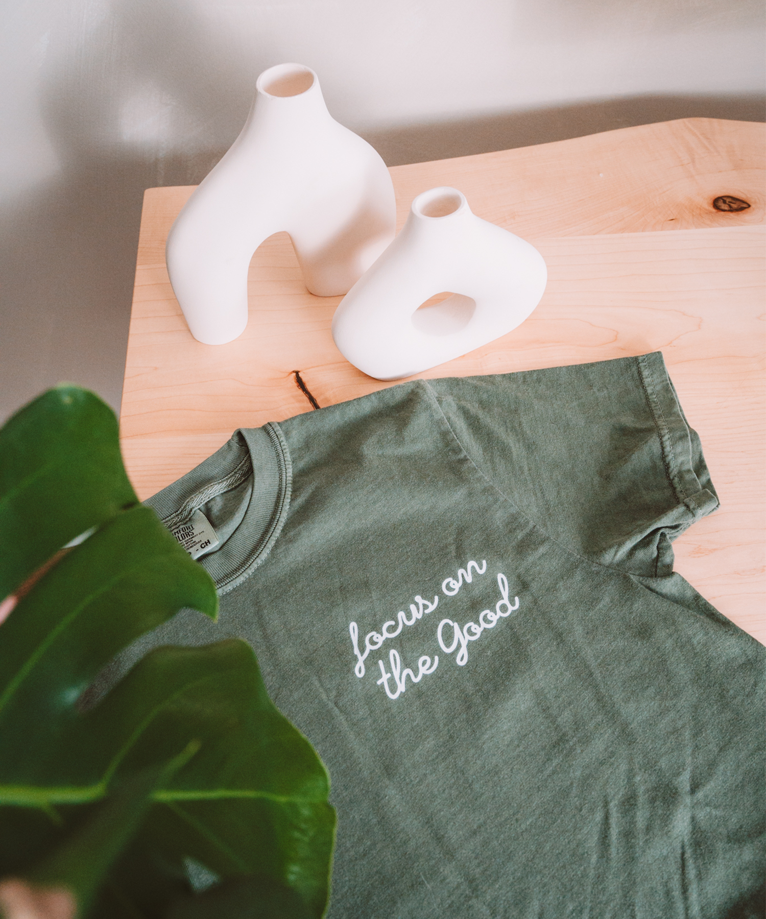 The "Focus on the Good" t-shirt in moss is laying on a desk with two asymmetrical vases in the front and a monstera leaf framing the front of the photo.