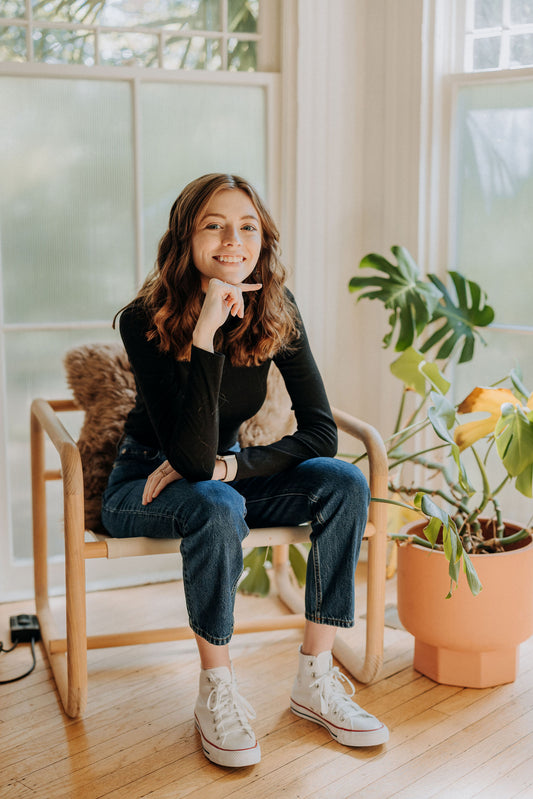 Kirstin from KDigitalStudio is sitting in a chair next to a Monstera, in front of large windows. She's wearing a black turtle neck, vintage blue jeans, and white high top converses to emulate the look of Steve Jobs.