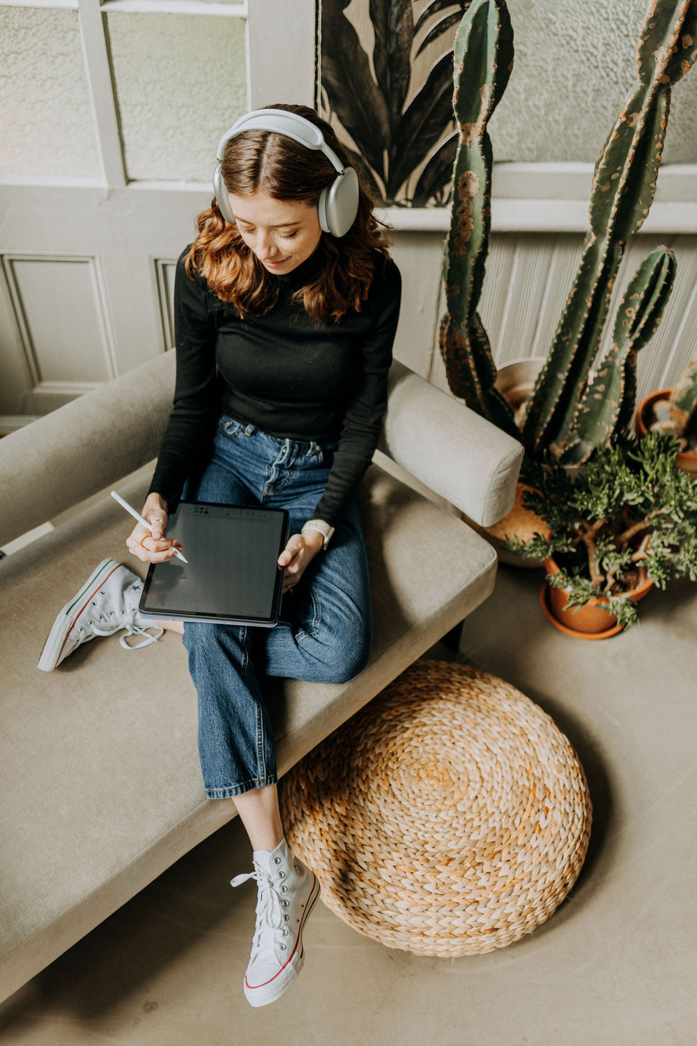Kirstin from KDigitalStudio is lounging on a couch while wearing Airpod Max headphones and writing on the iPad. She is surrounded by plants.