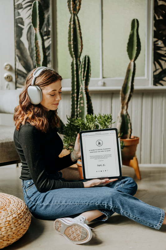 Kirstin from KDigitalStudio is sitting on the floor with her iPad and is wearing Airpods Max headphones. On her iPad is a guide for digital planning. She is surrounded by large cactus plants.