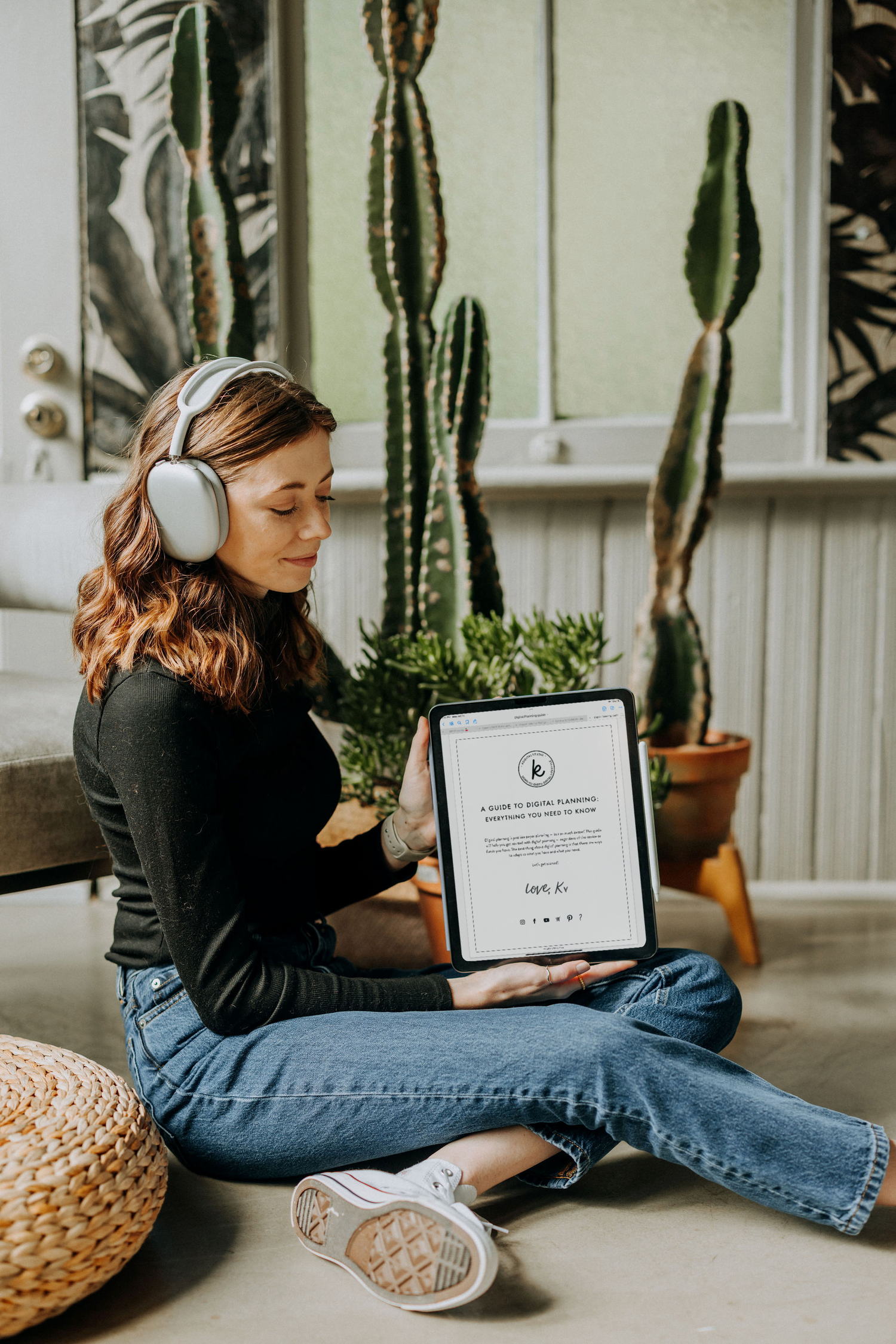 Kirstin from KDigitalStudio is sitting in front of large cactus plants, wearing headphones and holding her iPad, which shows a guide for digital planning.