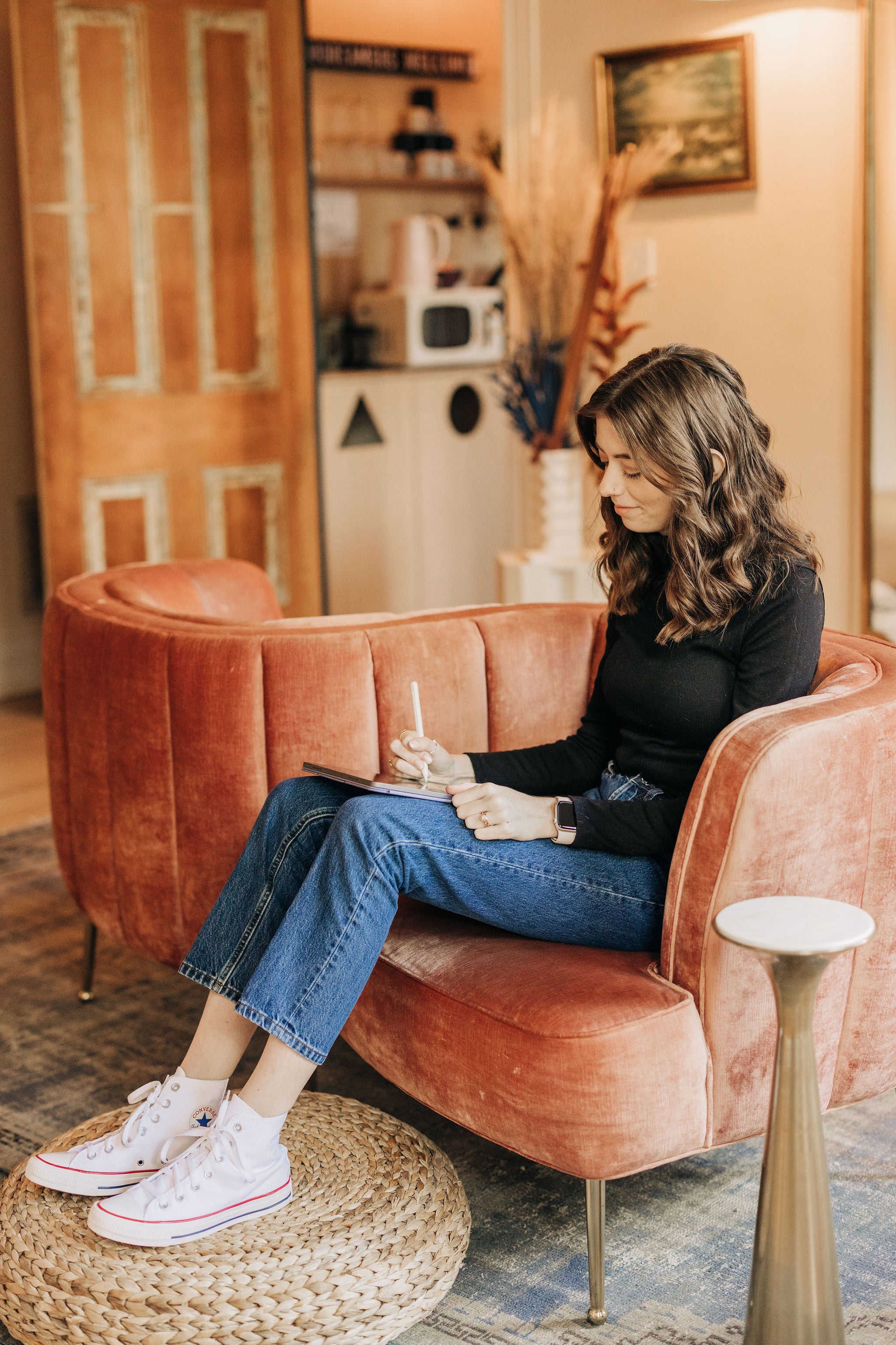 Kirstin from KDigitalStudio is sitting on a pink conversation couch. She has her feet propped up on a boho poof and has her iPad in her lap. She is wearing a black turtleneck, vintage blue jeans, and white Converse hightops.