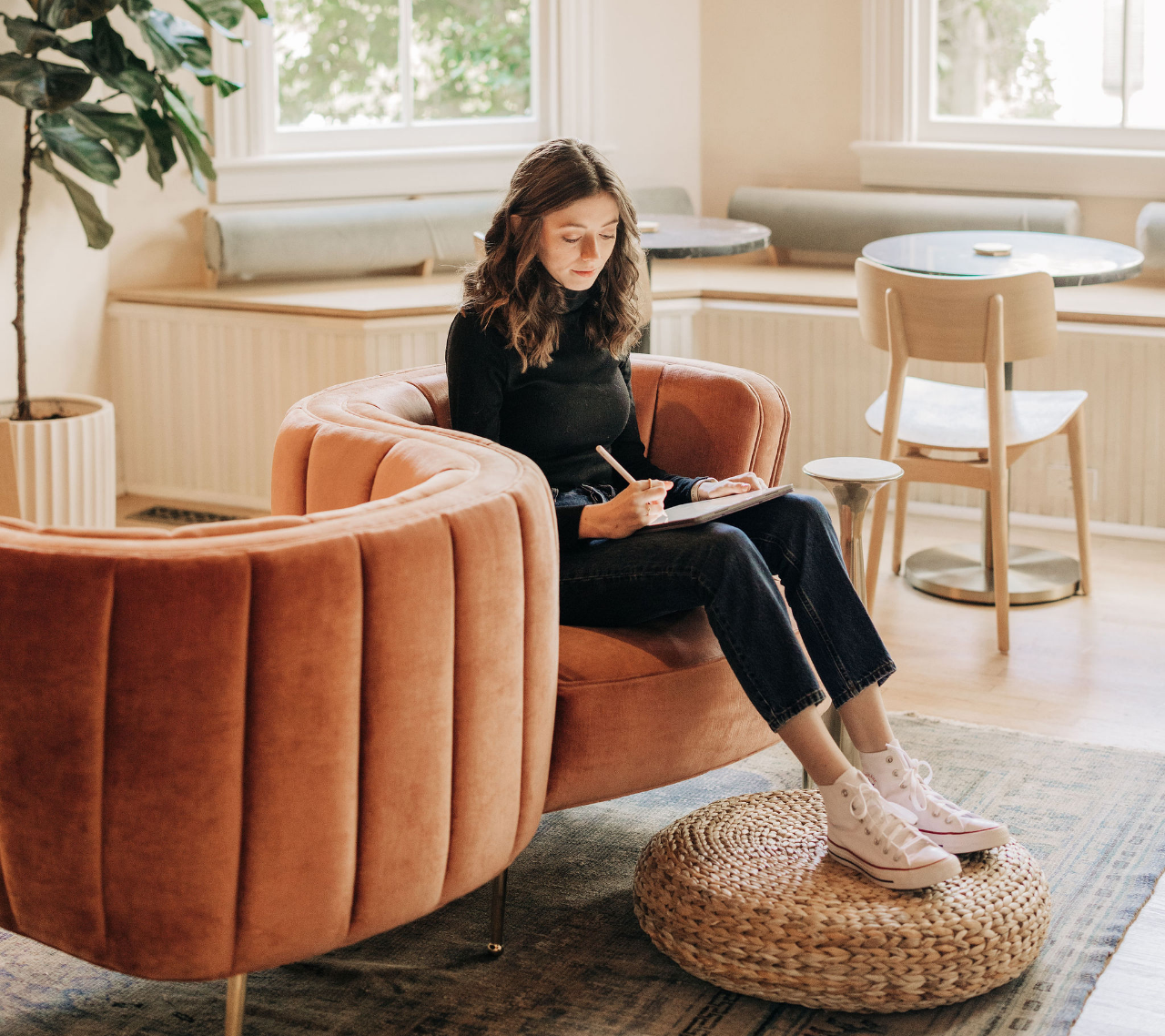 Kirstin from KDigitalStudio is sitting on a conversation couch with her feet propped up on a boho-style poof. She has her iPad in her lap and is writing on it with the Apple Pencil.