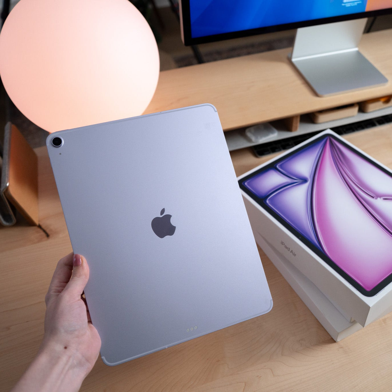 Person holding an Apple product on a desk with a box and monitor in the background