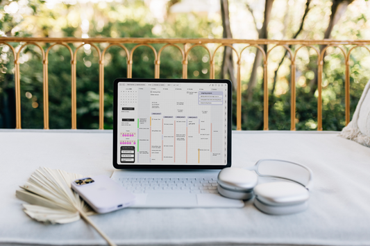 The Magic Keyboard is attached to the iPad. Sitting around the iPad are Airpods Max headphones, an iPhone, and a boho leaf fan. Featured on the iPad is a landscape weekly page of the Cyberry digital planner.